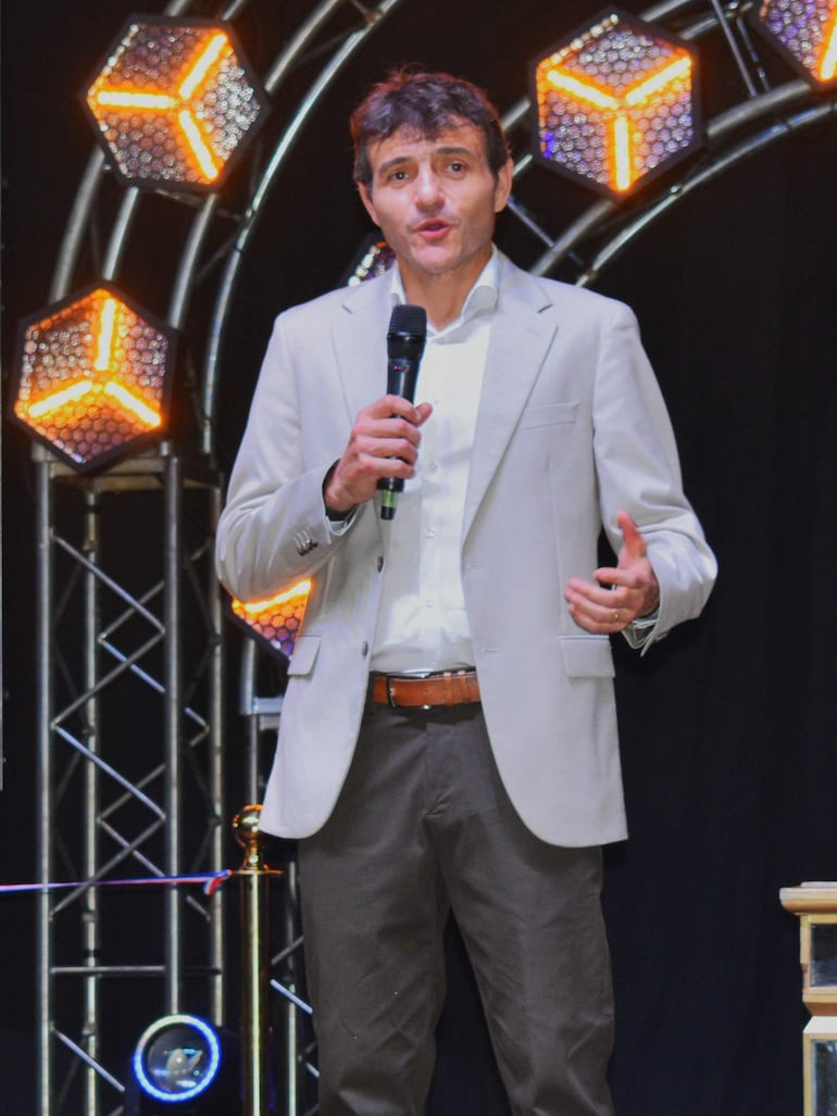 Hombre en traje claro y camisa blanca habla con micrófono, frente a un fondo de luces LED en un evento formal.