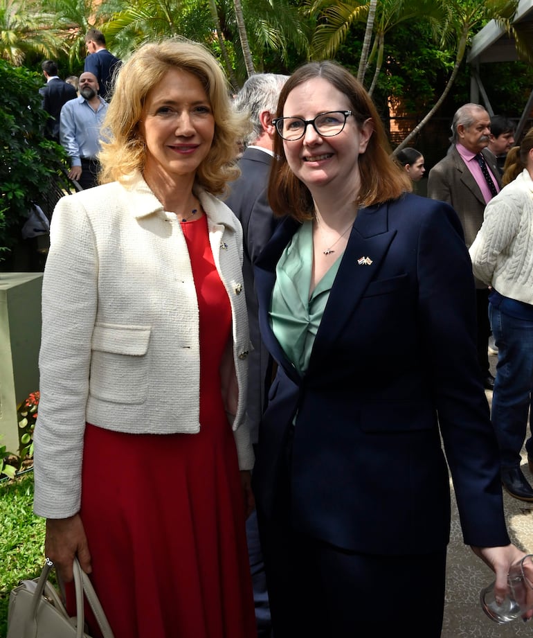 Mujeres sonrientes, una con vestido rojo y chaqueta blanca, y otra con traje oscuro y blusa verde, en un evento al aire libre.