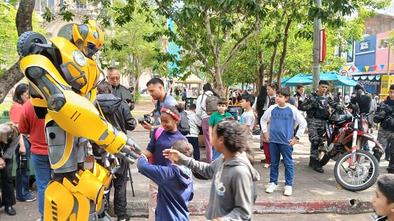 Grupo de niños rodea a un robot amarillo en un ambiente festivo, con adultos observando y decoraciones coloridas alrededor.