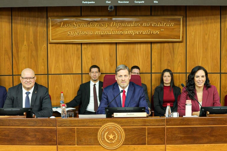 Un hombre con traje azul y corbata roja junto a una mujer con blazer burdeos, ambos sonrientes en un recinto legislativo.