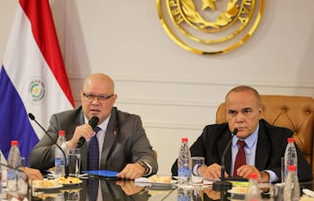 Óscar Lovera habla con seriedad, mientras otro hombre escucha atentamente en una reunión oficial con banderas de Paraguay al fondo.
