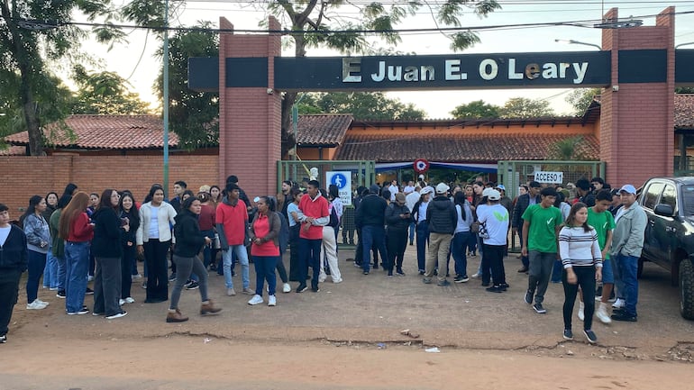 Grupo de jóvenes frente al edificio E Juan E. O'Leary, algunos en movimiento, vestidos de manera variada en un ambiente exterior.