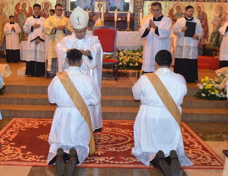 Obispo en mitra y báculo impone manos sobre Joel Ireneo Benítez Gayoso, vestido de túnica blanca, en un ambiente religioso.