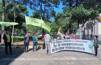 Un grupo de personas sostiene banderas verdes de OTEP-SN y un cartel en un parque de Asunción, mostrando expresiones de seriedad.