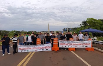 Un grupo de hombres con camisetas oscuras y un banner sobre el paso de camiones en un día nublado, con una noria al fondo.