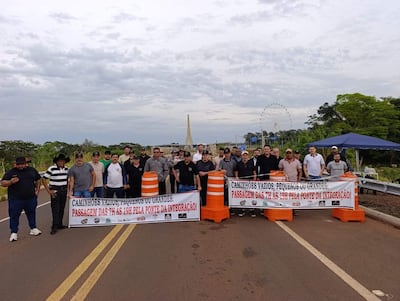 Un grupo de hombres con camisetas oscuras y un banner sobre el paso de camiones en un día nublado, con una noria al fondo.
