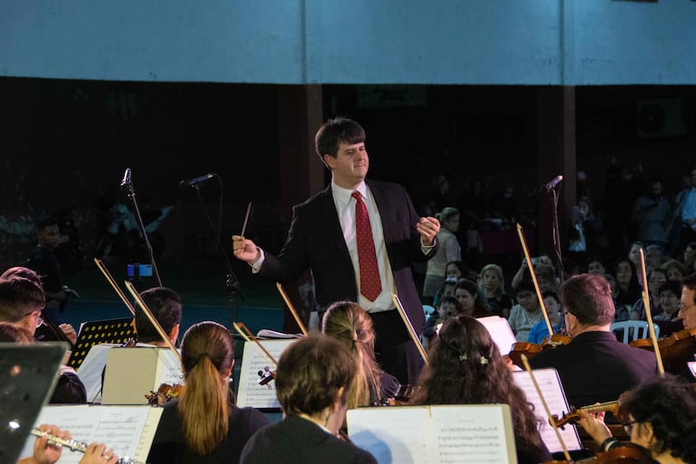 Director de orquesta con traje oscuro y corbata roja dirigiendo a músicos en recital al aire libre, público atento al fondo.