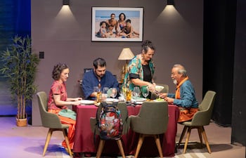 Mujer de blusa negra sirve comida al hombre mayor con suéter azul, en un comedor decorado con elementos familiares.