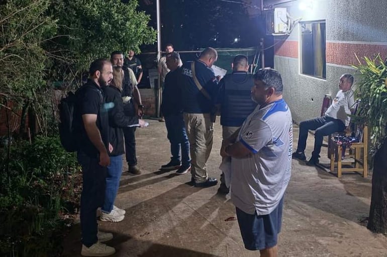 Grupo de ocho personas en la entrada de una vivienda, con un hombre en camiseta de fútbol blanco y azul, conversando animadamente.