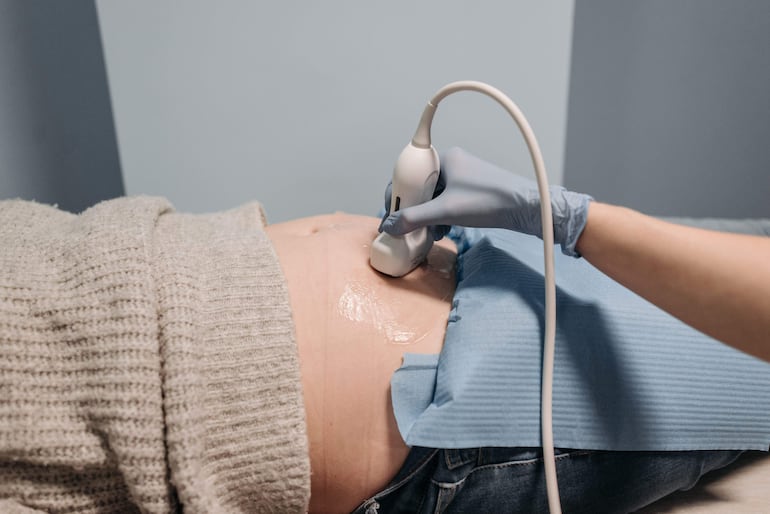 Mujer embarazada con abdomen expuesto y paño azul, mientras un médico con guantes sostiene un ultrasonido en un entorno clínico.