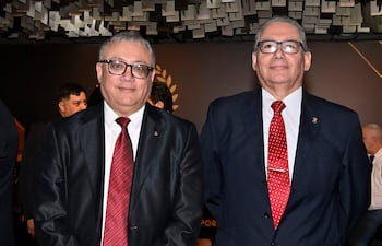 Dos hombres en trajes oscuros y corbatas rojas, con expresiones neutras, en un evento formal de fondo oscuro.