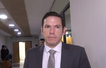 Alejandro Aguilera, vestido con traje oscuro y corbata de estampado, de pie en un pasillo, mientras otras personas observan atentamente.