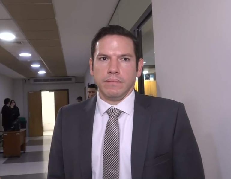 Alejandro Aguilera, vestido con traje oscuro y corbata de estampado, de pie en un pasillo, mientras otras personas observan atentamente.