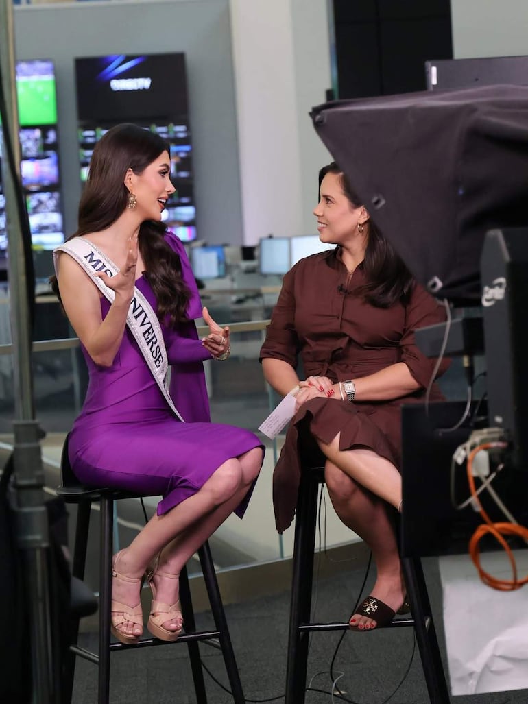 Gretha Matiauda en blusa marrón y participante de Miss Universe en vestido morado, sentadas en taburetes conversando en un estudio.