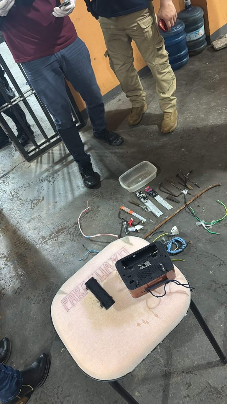 Dos hombres, uno con camiseta burdeos y guantes, y otro con camiseta negra, inspeccionan objetos en un taller desordenado.