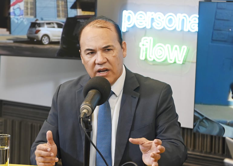 Hombre en traje oscuro y corbata azul habla en un estudio, con micrófono frente a él y un cartel iluminado de fondo.