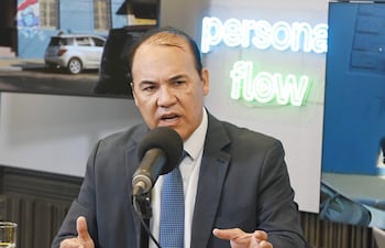 Hombre en traje oscuro y corbata azul habla en un estudio, con micrófono frente a él y un cartel iluminado de fondo.