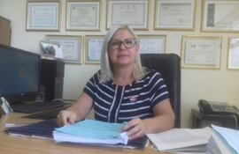 María Griselda Caballero, con cabello rubio y gafas, viste camiseta a rayas, sostiene documentos en su oficina ordenada con diplomas enmarcados.