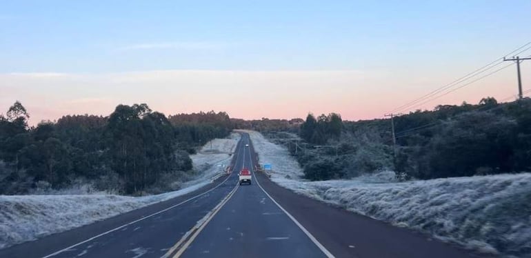Carretera helada en un entorno natural, sin personas, con un vehículo rojo circulando entre árboles y colinas.