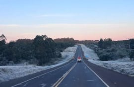 Carretera helada en un entorno natural, sin personas, con un vehículo rojo circulando entre árboles y colinas.