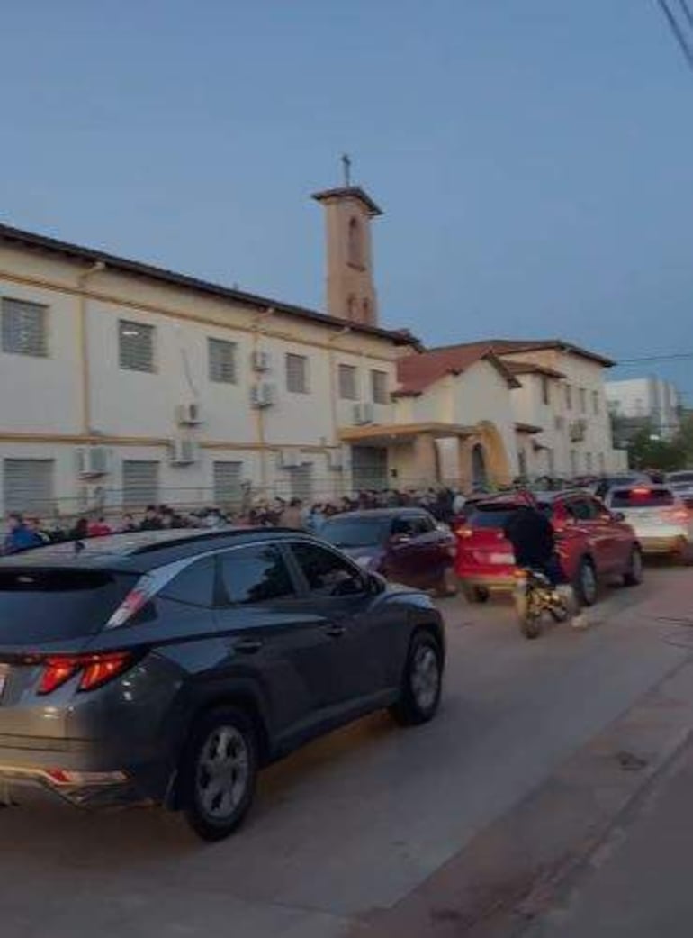 Personas en fila frente a un colegio, con vehículos alrededor y ambiente urbano en horas de la tarde.