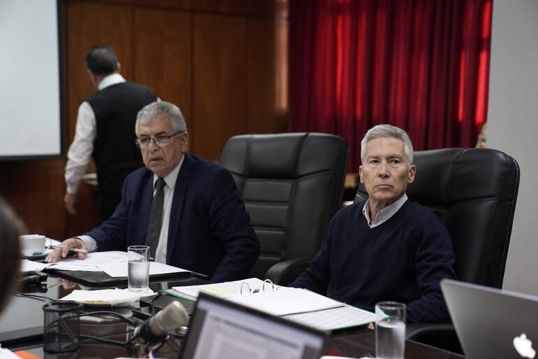 José Emilio Argaña Contreras en suéter oscuro y José Jara Rojas en traje oscuro con anteojos, en mesa de reunión formal.