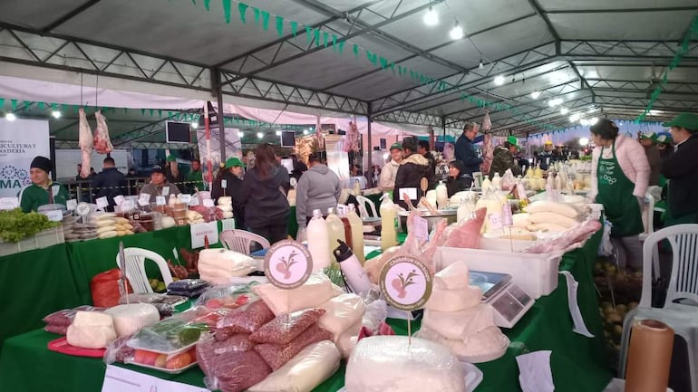 Personas adultas interactuando en un mercado al aire libre, rodeadas de puestos que ofrecen verduras, quesos y carnes, algunos vestidos con camisetas verdes.