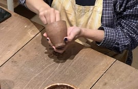 Dos artesanos trabajan en barro, uno modela una taza y el otro un plato, en un taller de cerámica con materiales diversos.