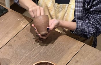 Dos artesanos trabajan en barro, uno modela una taza y el otro un plato, en un taller de cerámica con materiales diversos.