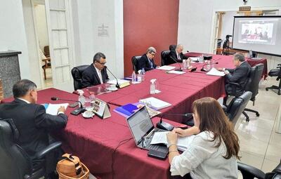 Seis participantes en reunión formal, vestimenta de traje, con mujer tomando notas en su computadora portátil.