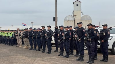 Policías uniformados en formación, con armas, en el estacionamiento de la Playa San José. Bandera de Paraguay al fondo.