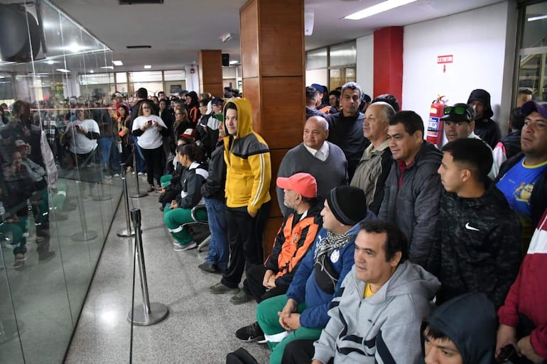 Grupo de funcionarios en sala de espera, mientras algunos miran expectantes. Predominan ropa en negro, verde y rojo, con un hoodie amarillo destacado.