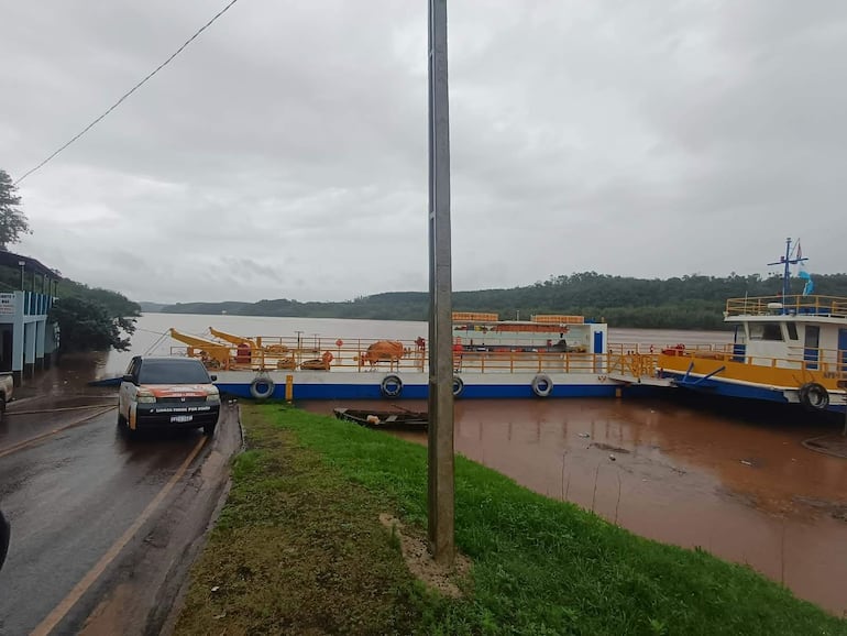 Ambiente nublado en puerto con edificio parcialmente inundado y ferry amarrado, vehículo blanco estacionado en carretera mojada.