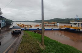 Ambiente nublado en puerto con edificio parcialmente inundado y ferry amarrado, vehículo blanco estacionado en carretera mojada.
