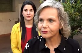 Lilian Samaniego con blusa oscura y cabello rizado, hablando seriamente, mientras otra mujer con blusa roja y abrigo amarillo escucha atentamente.