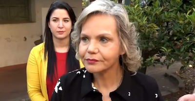 Lilian Samaniego con blusa oscura y cabello rizado, hablando seriamente, mientras otra mujer con blusa roja y abrigo amarillo escucha atentamente.
