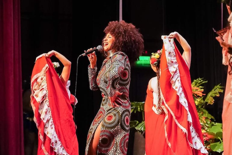 Concha Bernal, con vestido colorido y micrófono, canta enérgicamente rodeada de bailarinas con flores en el cabello en un ambiente teatral.