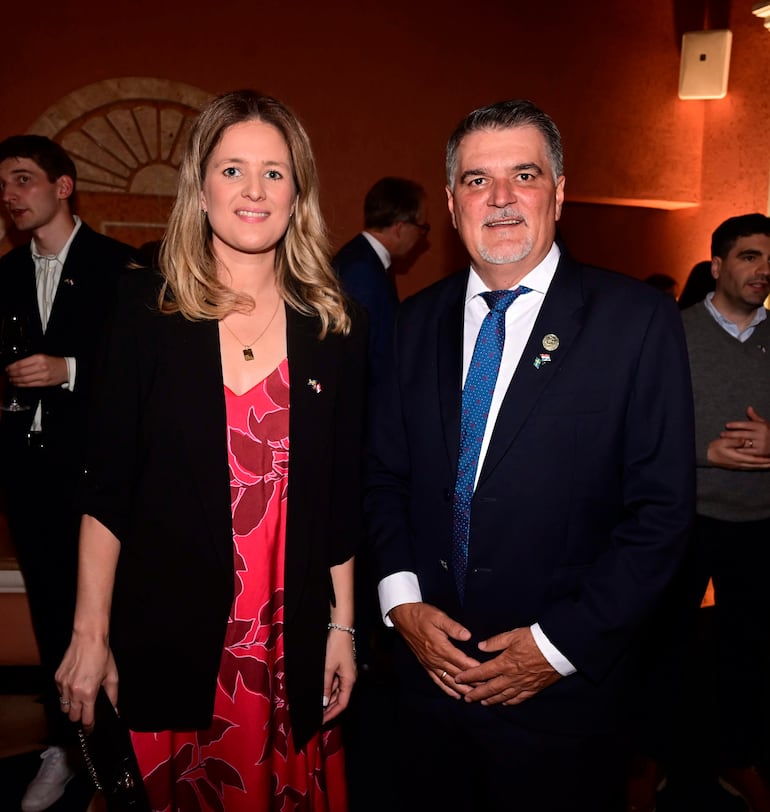 Mujer de cabello rubio y vestido floral rojo, junto a hombre canoso con traje oscuro en ambiente elegante.
