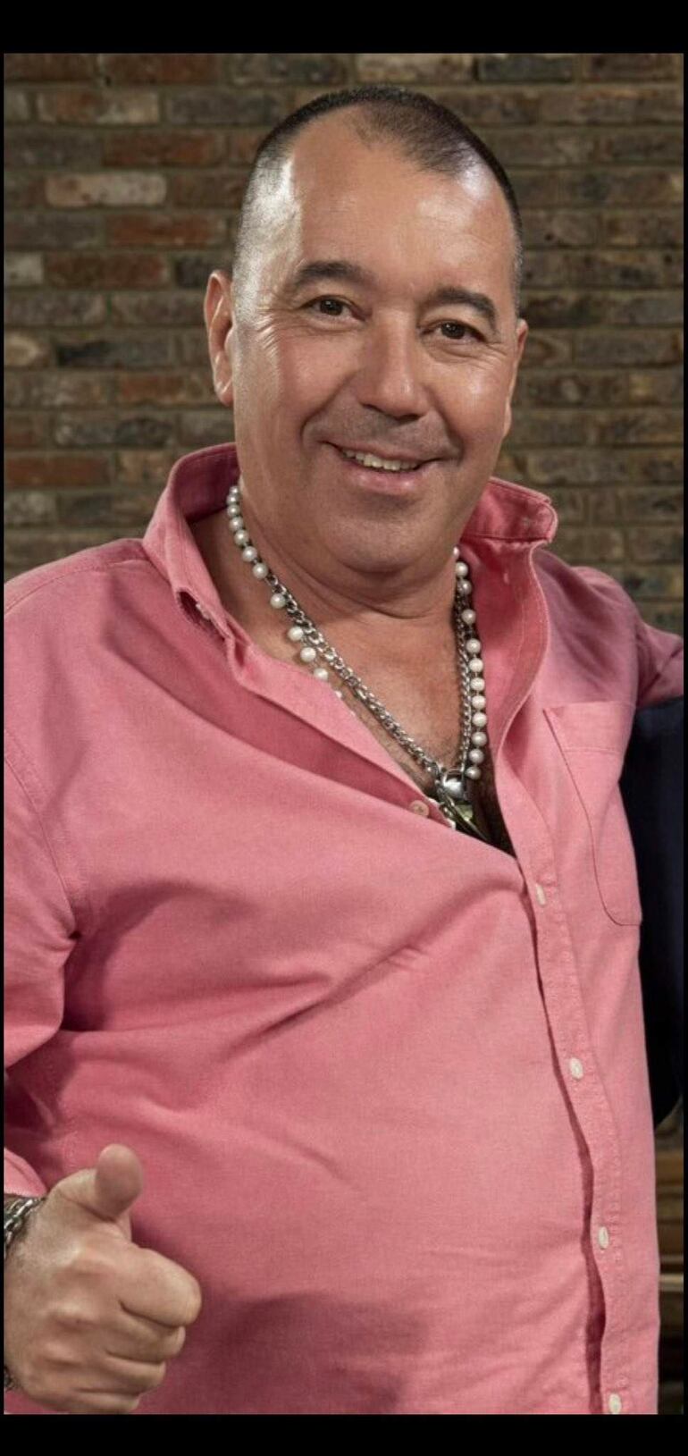 Hombre sonriente en camisa rosa clara, con collares de cuentas, hace un gesto de pulgar hacia arriba en ambiente casual.
