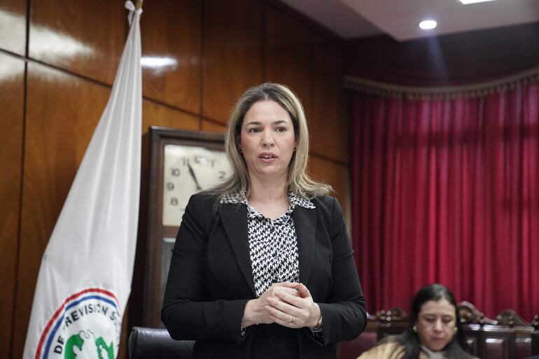 Mujer de cabello largo y rubio en blazer negro habla ante un salón con fondo de madera y cortinas rojas.