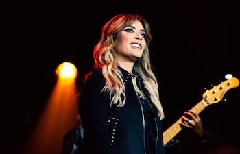 Kany García sonríe en el escenario con chaqueta negra mientras sostiene un micrófono. Músico detrás con un bajo.