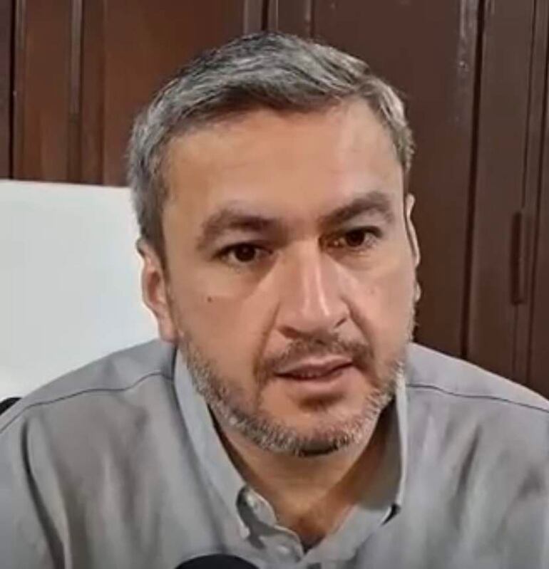 Hombre de mediana edad con barba y camisa clara, hablando frente a un micrófono en un entorno formal de sala de reuniones.