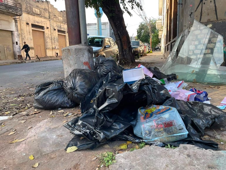 Hombre con ropa oscura y mochila camina entre bolsas de basura negra acumuladas en una calle descuidada.