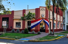Edificio municipal en San Juan Bautista con colores terracota y crema, adornado por una bandera paraguaya y un mural a la izquierda.