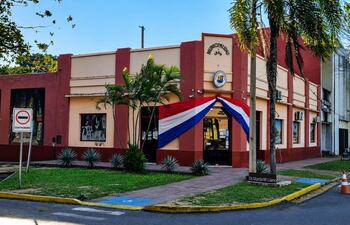 Edificio municipal en San Juan Bautista con colores terracota y crema, adornado por una bandera paraguaya y un mural a la izquierda.
