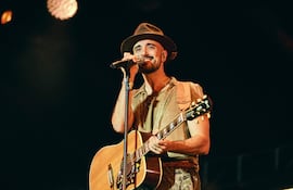 Abel Pintos sonriente, vestido con camisa clara y pantalones camuflados, sostiene guitarra y micrófono en un escenario iluminado.