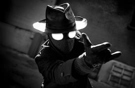 Figura con traje negro y sombrero, brazo extendido en un entorno urbano sombrío y en blanco y negro.