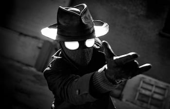 Figura con traje negro y sombrero, brazo extendido en un entorno urbano sombrío y en blanco y negro.