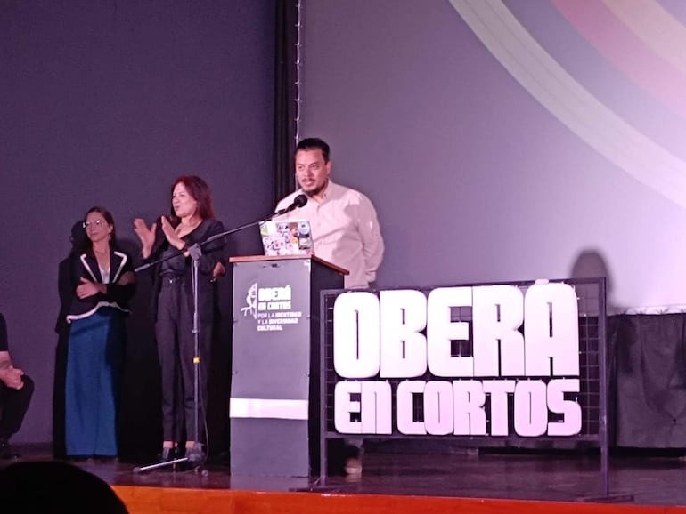 Hombre en camisa clara habla en micrófono, con panel de 'OBERÁ EN CORTOS', dos mujeres aplauden a su lado.