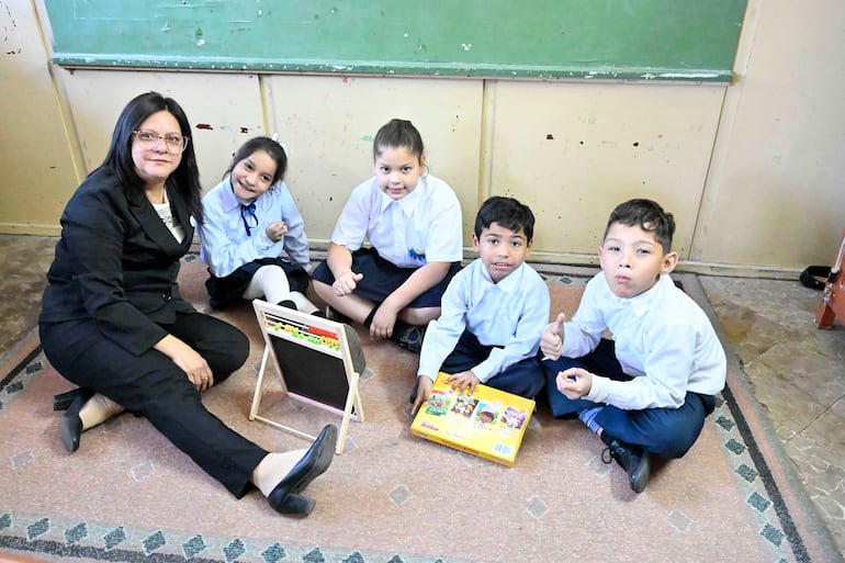 Mujer con traje negro interactúa con niños en un aula, con pizarra verde y juegos educativos.